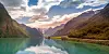 Wide fjord landscape with mountains, autumn trees and a reflective lake under a colourful sunset sky.