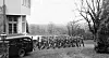 A number of German soldiers march away from Skaugum past a building and a parked car.