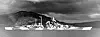A black-and-white photograph of a warship moored at a quay with mountains and an industrial area in the background.