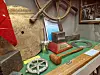 A historical maritime exhibition with a ship’s wheel, ropes, metal objects, and models displayed on a green table.