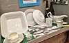 White tableware, cups, cutlery, and bottles displayed on a bench.