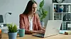 Woman using a laptop at a desk in a bright home office with plants and shelves.