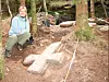 Irene Baug sits beside a large stone cross lying on the ground. In the background are trees and the waste heap from the old millstone quarry.