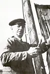 A black-and-white photo of a man in work clothes standing beside a post outdoors.