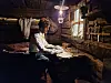 A person works at a wooden table in a dim rustic workshop with shelves of tools and supplies.