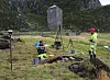 People working in a grassy field beside a stone tower, with tripods and equipment nearby.