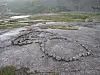 Circular stone formations on a grey rocky hillside with patches of grass and distant mountains.