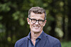 Smilende mann med briller og blå skjorte foran grønn, uskarp bakgrunn.