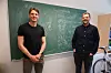Two men stand in a classroom beside a chalkboard covered with handwritten model equations.
