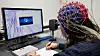 Participant in an EEG cap writes beside a computer monitor during a brain-response study.