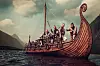 Vikings aboard a carved dragon-headed longship sailing on dark water beneath cloudy skies.