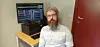 Bearded man seated beside a computer monitor in an office with a red door.