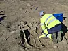 An archaeologist kneels and excavates a grave – we can see three skeletons in the soil.