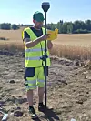 Archaeologist Aksel Haavik stands in the field writing in a small yellow notebook. He is wearing a high-visibility vest and yellow shorts.