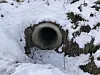 Snow-covered circular culvert opening in a railway embankment, surrounded by exposed earth and frost.