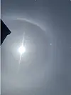 The sun shines through a thin layer of cloud, with a faint ring of light around it and the dark edge of a building on the left.