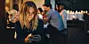 Woman looking at her phone while people socialise at a candlelit gathering.