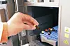 A hand places a small sample into laboratory equipment beside a rack of coloured-capped tubes.