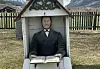 A small, chapel-shaped headstone featuring a sculpture and an open book, situated on a lawn in front of mountains and a fence.