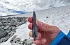Image of a hand holding an ancient arrowhead. In the background, a snow patch and numerous rocks can be seen.
