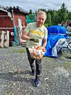 Man holding a basket of eggs in a gravel farmyard beside a red timber building.
