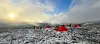Tents in the mountains, several of them red and some brown. The sun is rising, and the sky is blue with some cloud cover. Fresh snow covers the ground.