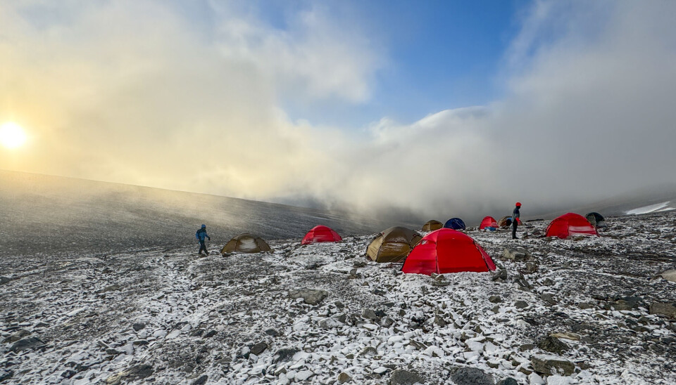 Tents in the mountains, several of them red and some brown. The sun is rising, and the sky is blue with some cloud cover. Fresh snow covers the ground.