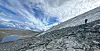 Dramatic mountain landscape. An ice patch on the right, a small lake on the left. An archaeologist walks down the mountainside.