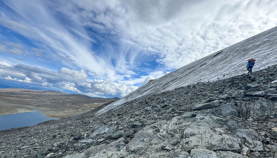 Dramatic mountain landscape. An ice patch on the right, a small lake on the left. An archaeologist walks down the mountainside.