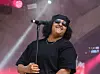 Musti sings into a microphone on a dimly lit stage, wearing sunglasses and black clothing.