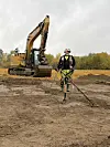 An excavator in the background in a field. A metal detectorist stands in the middle of the picture, searching the soil with a metal detector.