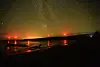 Offshore wind turbines with red aviation lights reflected on dark water beneath a starry night sky.