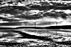 Black-and-white coastal landscape with three wind turbines on the horizon beneath dramatic clouds.