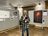 Neil Gordon Davey stands in a gallery beside framed exhibition artwork with a glowing red symbol.