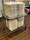 A metal trolley loaded with stacked covered food trays in a commercial kitchen.