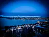 Winter view of Tromsø harbour, snowy rooftops, city lights and mountains under a blue evening sky.
