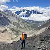 Lotta in an orange jacket and blue backpack on a rocky mountain trail beneath snow-capped peaks.