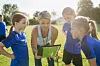 A coach shows a tablet to four young athletes on a grass field.