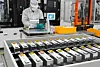 Worker in protective clothing oversees rows of battery cells on an industrial production line.
