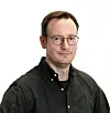 Man wearing glasses and a dark shirt, photographed against a light background.