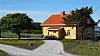 Yellow rural house with a red roof, gravel driveway and trees under a blue sky.