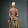 Weathered painted wooden figure seated on a carved chair against a dark background.