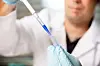 Gloved laboratory worker transferring blue liquid with a pipette into a small vessel.