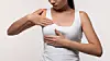 Woman in a white top examining her breast with her hands against a light background.