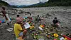 Relatives perform rituals among flowers and food offerings on a gravel riverbank beside the Trishuli River in Nepal.