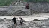 Two people cross a rocky, flood-affected river while a zipline basket hangs above the water in Nepal.