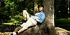 Man sitting against a tree in a sunlit woodland, with one knee raised.
