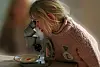 Young student in a pink jumper looks through a microscope beside a specimen dish.