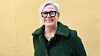 Smiling woman in glasses and a dark green coat against a yellow wall