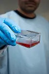 Blue-gloved hand holding a clear laboratory container with red gel against a blurred lab coat.